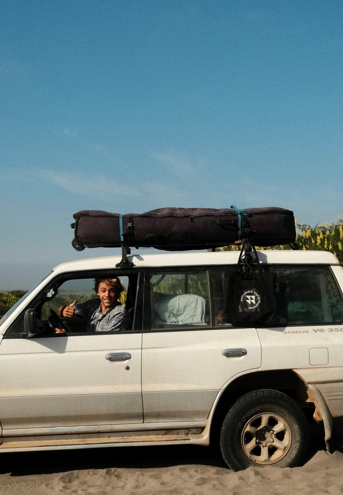 White SUV with luggage on top, person inside giving a thumbs-up, clear blue sky